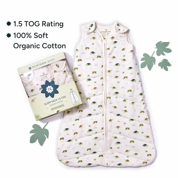 Nintara Baby sleep sack laid flat beside its gift box packaging. Text highlights 1.5 TOG rating and 100% soft organic cotton. Decorative green leaves styled around the product on a white surface.