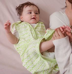 Close-up of a baby lying on a bed wearing a sleeveless green and white patterned sleep sack with a front zipper. The infant is interacting with an adult whose hand gently holds the baby’s foot. The baby has light skin and dark hair, and the background shows soft bedding in muted tones, emphasizing comfort, safe sleepwear, and bonding time between parent and child.