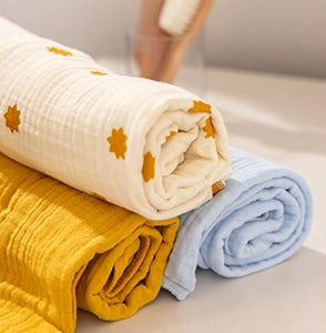 Image of three soft muslin baby blankets neatly rolled and stacked on a white surface. The top blanket is cream-colored with mustard yellow starburst prints, the middle is mustard yellow, and the bottom is light blue. A blurred background with a glass container holding a baby brush is visible, highlighting the cozy and premium texture of the blankets designed for infants.