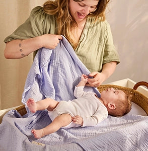 Smiling woman with light brown hair wearing a green blouse playfully holds a soft light blue muslin blanket above a baby lying in a woven changing basket. The baby, dressed in a white onesie, kicks happily while gazing up. The room is warmly lit, with neutral-toned curtains in the background, highlighting bonding moments during diaper changes and the comfort of breathable fabrics.