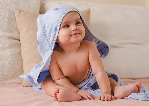 Adorable baby sitting upright on a soft surface, wrapped in a light blue muslin hooded towel with blue starburst prints. The hood gently covers part of the baby’s head, leaving the face visible as the baby smiles with curiosity. A beige cushion rests in the background, creating a cozy, homelike setting that emphasizes comfort, warmth, and post-bath care for little ones.