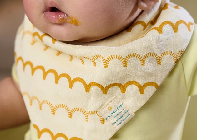 Close-up of a baby wearing a yellow shirt and a cream-colored bib featuring a mustard yellow arch pattern. A small amount of food can be seen on the baby's chin, likely from the baby’s mealtime. The Nintara Baby logo is visible on the bib's label, adding a touch of authenticity. The background is softly blurred, emphasizing the bib’s absorbent and stylish design, perfect for mealtime messes.