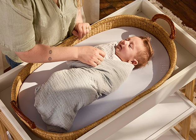 A peaceful moment with a baby wrapped in a soft grey swaddle blanket lying in a wicker basket. The baby’s face is calm and content, with closed eyes. A woman’s hand is gently adjusting the swaddle, ensuring comfort. The natural light filtering through the room enhances the soothing ambiance, creating a scene that reflects safety, warmth, and nurturing care for newborns.
