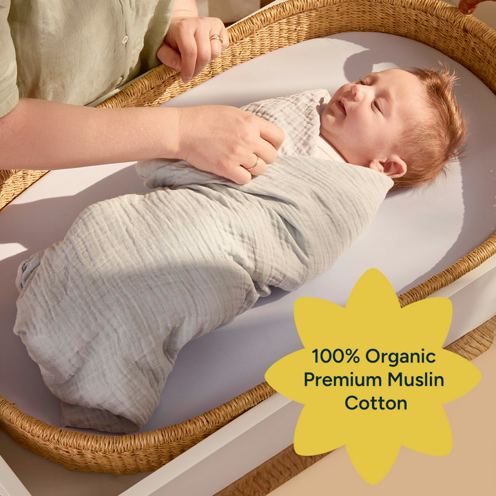 A gentle moment shows a parent adjusting the swaddle on a baby, who is wrapped in a soft, light green organic muslin cotton fabric. The image conveys warmth and care, as the baby looks relaxed, lying on a soft surface. The swaddle’s breathable material ensures comfort while maintaining its durability, with the text overlay promoting its 100% organic cotton quality.