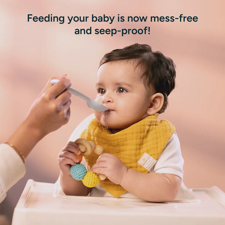 A soft, calming image of a baby being fed while wearing a Nintara Baby bib. The bib is made from soft, organic cotton and is designed to be mess-free and seep-proof. The bib's mustard yellow color adds a warm touch, while the gentle texture of the fabric enhances its comfort and absorbency. The background is softly blurred, focusing on the baby’s peaceful expression as they enjoy their meal, demonstrating the bib’s practical design and comfort during feeding.