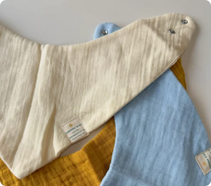A close-up of three baby bibs. The first bib is a light beige color, the second one is a soft blue, and the third is mustard yellow. All three bibs are made from a soft, breathable fabric with snap buttons for easy closure. Each bib features a small Nintara Baby label, adding a branded touch. The textures and colors reflect comfort and practicality, ensuring they are gentle on a baby’s skin.