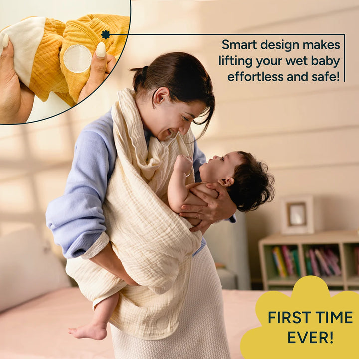 A mom smiling down at an infant while lifting them from a bath using a cream-colored muslin baby towel wrap worn over her shoulder like a sling. The soft, crinkled towel forms a secure pouch around the baby's lower body; the mom supports the child with one arm and steadies the pouch with the other. She wears a pastel blue sweater and a textured off-white skirt. The image demonstrates hands-free, safer post-bath handling enabled by the towel's shoulder-worn, fastened design.