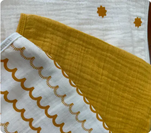 A set of three muslin cloths folded neatly. The top cloth is white with a pattern of small, scattered orange stars. The middle cloth is yellow with a scalloped design in a deep orange color, and the bottom cloth is a solid mustard yellow. The textures of the fabrics are visible, showing their softness and high-quality finish, perfect for babies. The design elements are gentle and eye-catching, ideal for baby accessories.
