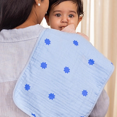 A baby is held by their mother, who is wrapped in a soft blue muslin cloth adorned with blue star patterns. The baby gazes up with a joyful expression, showing their curiosity and excitement. The mother is dressed in a light grey shirt, with warm, soft lighting highlighting the cozy atmosphere.
