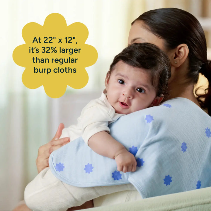 A lifestyle product image shows a mother holding her baby upright against her shoulder, demonstrating the size and comfort of a large baby burp cloth. The cloth, draped over the mother's shoulder, is light blue with a repeating pattern of darker blue floral star motifs. The baby, dressed in a cream outfit, rests comfortably on the cloth, facing the camera with a curious expression. - The scene highlights the product's generous size, softness, and practicality for everyday use.