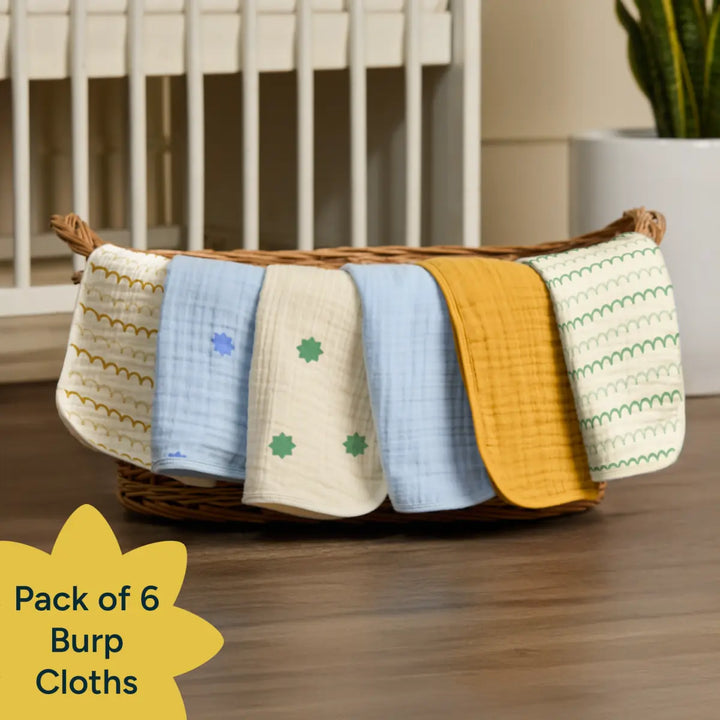 A cozy nursery setting shows basket with a neatly arranged pack of six colorful burp cloths. Each cloth drapes over the edge of the basket to display its design: one with golden scalloped patterns on white, one in soft blue with small floral prints, one in cream with green motifs, one in solid mustard yellow, and one in cream with green arches. The fabrics appear soft and textured In the bottom left corner, a yellow flower-shaped graphic contains the text: "Pack of 6 Burp Cloths.
