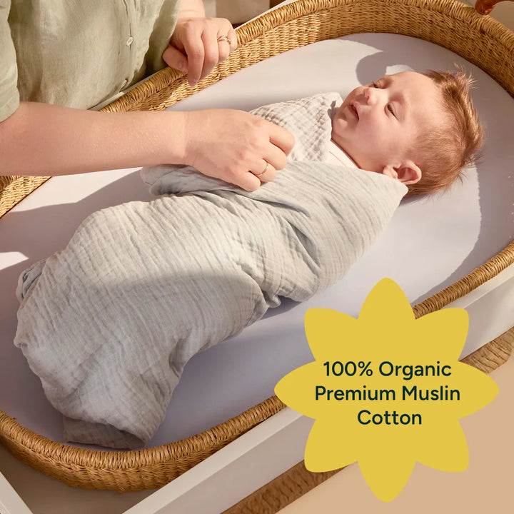 A gentle moment shows a parent adjusting the swaddle on a baby, who is wrapped in a soft, light green organic muslin cotton fabric. The image conveys warmth and care, as the baby looks relaxed, lying on a soft surface. The swaddle’s breathable material ensures comfort while maintaining its durability, with the text overlay promoting its 100% organic cotton quality.