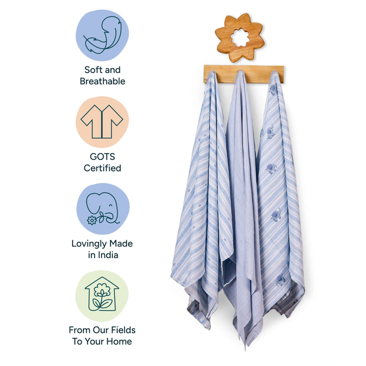 Three baby blankets are elegantly hanging on a wooden rack with a flower-shaped hook. Each blanket is made from soft, breathable fabric with light blue and white striped designs, featuring subtle prints of small animals. To the left of the image, icons with pastel backgrounds highlight the key features: soft, GOTS certified, lovingly made in India, and sourced directly from fields to your home. These attributes emphasize the product's eco-friendly and sustainable nature