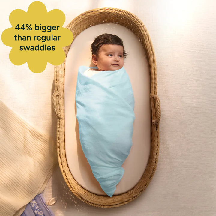 A baby rests peacefully in a cozy basket, wrapped in a soft blue organic cotton swaddle. The swaddle is 44% bigger than regular swaddles, providing ample space and comfort for the baby. Made from 100% organic cotton, it is gentle and breathable, offering a superior alternative to cotton-blend swaddles that often lose their softness.