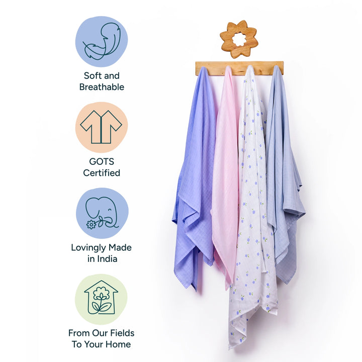 A set of four soft, pastel-colored organic cotton swaddles hangs neatly on a wooden rack, with a simple floral-shaped hook at the top. The swaddles are in varying shades of purple, pink, and blue, with delicate patterns. To the left of the swaddles are circular icons, each highlighting the key features of the product: soft, GOTS certified, lovingly made in India, and a statement of sustainability from the fields to your home.