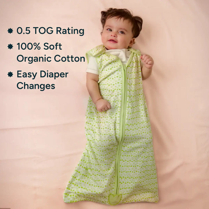 A smiling baby is lying on a soft pink backdrop, dressed in a green sleep sack with white polka dots. The sleep sack is made from 100% organic cotton, offering comfort for the baby. The image includes text features highlighting the product's 0.5 TOG rating for warmth, easy diaper changes with a zipper, and soft, breathable organic cotton, ensuring a cozy and safe sleep environment for babies.