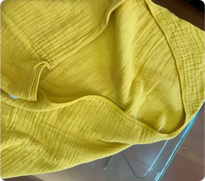 A close-up of a bright yellow muslin cloth, showing its soft texture. The fabric is gently draped, revealing the smooth and slightly wrinkled finish, highlighting its breathable and lightweight nature. This cloth would be ideal for use as a baby swaddle, blanket, or burp cloth, offering both style and comfort for infants.