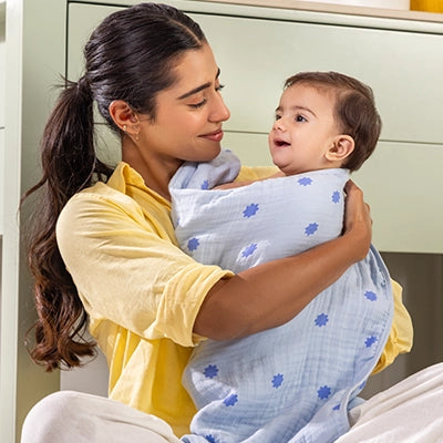 A smiling mother holds a baby wrapped in a light blue towel with small floral patterns. The baby looks up at the mother with a content expression. The background has a minimalist, modern nursery vibe with a few colorful toys, emphasizing warmth and comfort.