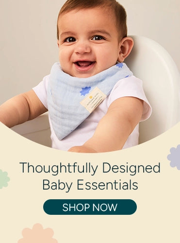 A smiling baby wearing a light blue bandana bib with floral designs, sitting in a highchair. The background features soft pastel flowers. The text 'Thoughtfully Designed Baby Essentials' is displayed next to the baby, with a call-to-action button saying 'SHOP NOW' in teal. The baby is enjoying the moment, adding to the friendly and nurturing vibe of the product image.