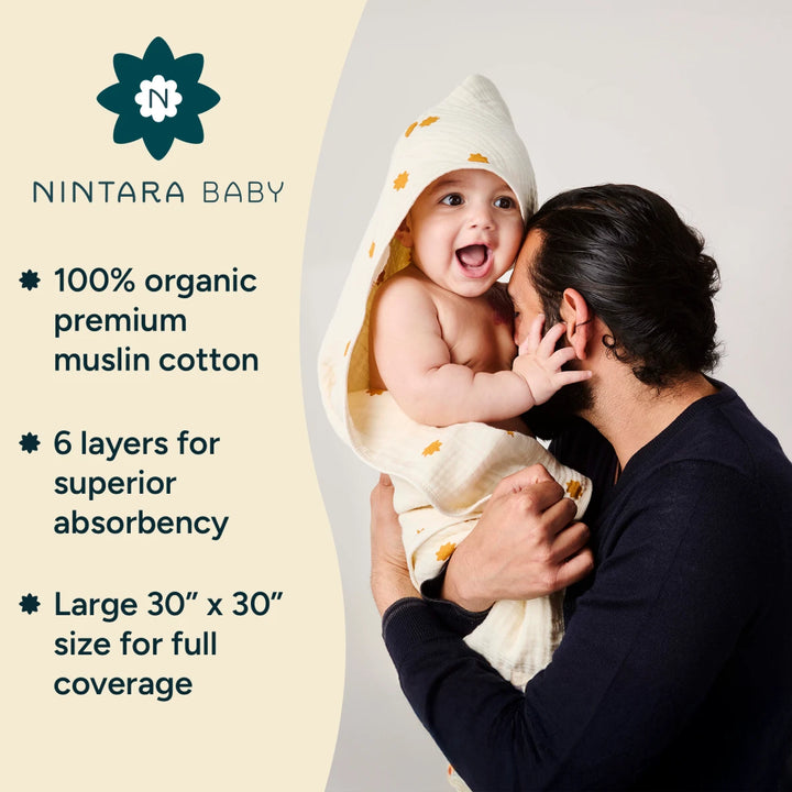 A father lovingly holds a smiling baby wrapped in a cream-colored hooded muslin towel adorned with small yellow prints. The baby's hood is draped over the head while the rest of the towel provides full coverage around the body. The father, wearing a dark shirt, gently kisses the baby's cheek, creating a warm and affectionate scene. - The Nintara Baby logo with a flower-shaped icon and stylized "N- appears above the text.