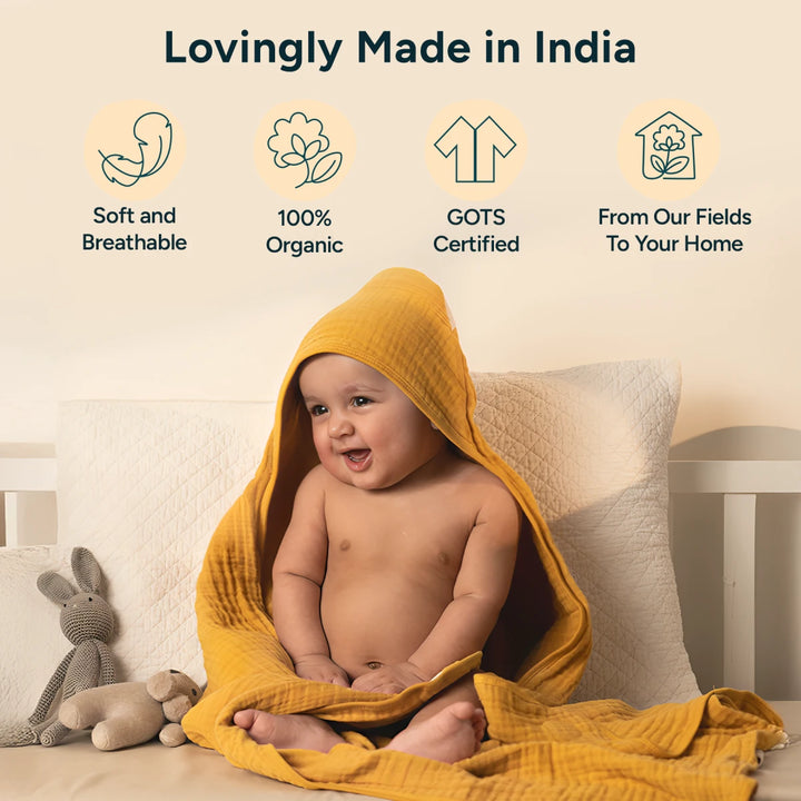 A cheerful baby sits on a white cushioned bench, draped in a mustard-yellow hooded muslin towel that wraps loosely around their body. The baby smiles joyfully, looking slightly to the side, with a hood covering the head and fabric pooling softly around the legs. On the left, text at the top reads "Lovingly Made in India,- accompanied by four icon illustrations. The first icon shows a curled fetus shape with the words "soft.