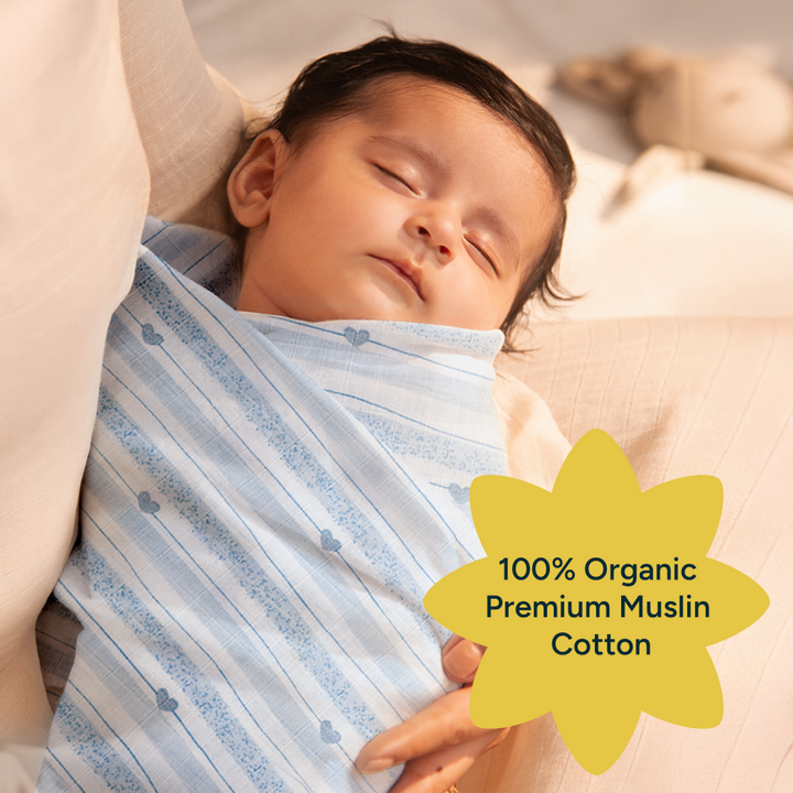 A peaceful baby, sound asleep, is wrapped in a soft blue swaddle with faint heart-shaped patterns. The gentle light from the surroundings accentuates the baby's calmness. The image is adorned with a bright yellow starburst graphic, highlighting the key feature of the swaddle: 100% organic premium muslin cotton. This visual reinforces the importance of organic materials for sensitive baby skin and assures parents of the product's softness, safety, and comfort for their little ones.