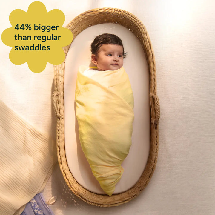 A smiling baby, wrapped in an extra-large yellow swaddle, resting in a bassinet. The image emphasizes the swaddle’s larger size compared to regular swaddles, making it easier for parents to wrap and use as the baby grows. A star-shaped label highlights the swaddle being "44% bigger than regular swaddles," offering added comfort and longevity. The baby’s expression reflects contentment, underscoring the swaddle’s gentle feel.