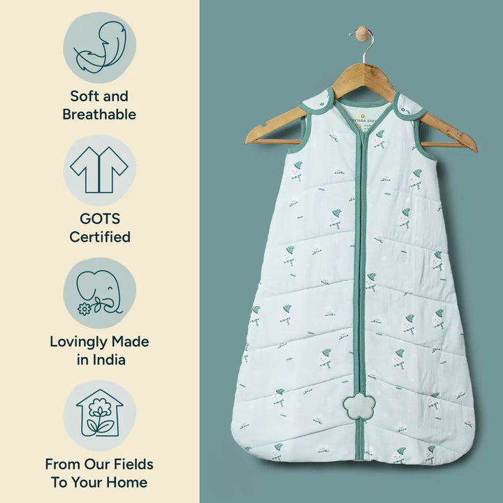 A beautiful product shot of the Nintara Baby sleep sack hanging on a wooden hanger. The sleep sack, made from 100% organic cotton, is shown against a teal-colored background that enhances its soft, pastel green tone. Next to the sleep sack are three icons: one indicating its softness and breathability, another confirming its GOTS certification, and a third showcasing that it is lovingly made in India.
