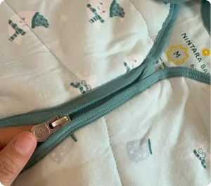 A close-up of a baby sleep sack with a light blue and white design featuring umbrellas. The zipper of the sack is visible, showing a smooth glide feature, and the Nintara Baby logo is also visible on the fabric. The soft, quilted design adds comfort and warmth, making it a perfect sleepwear choice for babies. The zipper's secure fastening ensures ease of use for parents.