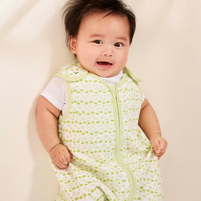 A cheerful baby standing and smiling with bright eyes. The baby is dressed in a white T-shirt and a light green sleep sack with a dotted pattern. Their hair is short and tousled, giving them an adorable, playful look. The background is a soft, neutral color, complementing the baby’s happy expression.