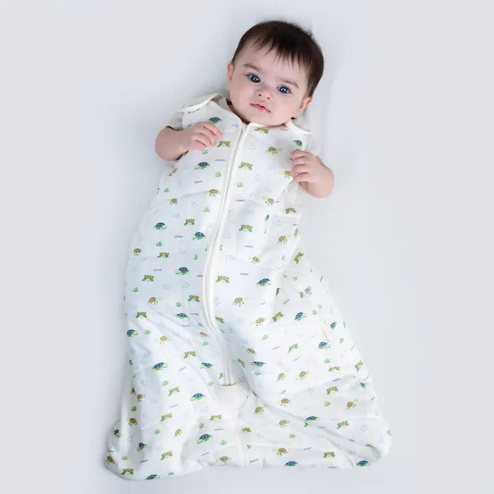 Baby on white cotton in Nintara Baby Snuggle Squad 1.5 TOG sleep bag. White fabric with colorful sea turtle, green seaweed, and blue wave print, cream piping trim, and shoulder snap closures.