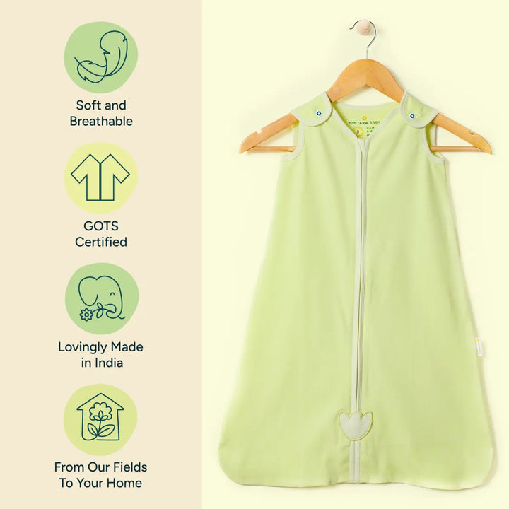 A pale green sleeveless baby sleep sack with a central zipper is displayed hanging on a wooden hanger against a cream background. The fabric is soft, lightweight, and gently structured, with rounded edges and a stitched heart detail near the bottom of the zipper. To the left of the image, four icons with text describe the product's features. The first icon shows a curled fetus shape with "soft.