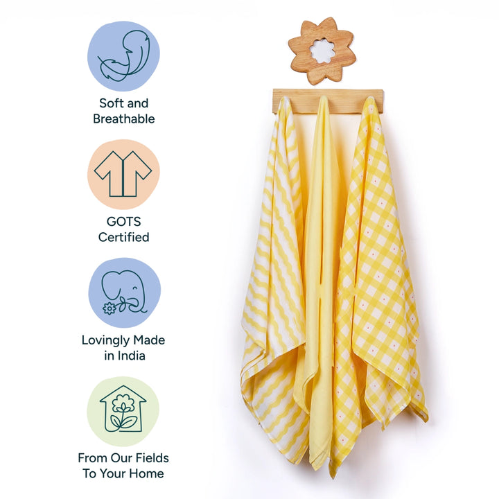 This image displays a set of three Nintara Baby muslin swaddles hanging on a wooden rack, with a wooden flower shape accent above them. Each swaddle is patterned in different designs such as stripes and checks in soft yellow tones. Beside the swaddles, icons highlight the product's key attributes: softness and breathability, GOTS certification, a commitment to being lovingly made in India, and the promise of quality materials sourced from the fields directly to homes.