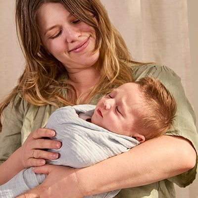 A loving mother cradling her newborn baby wrapped in a soft, light blue swaddle, gazing down affectionately. The mother’s joyful smile radiates warmth as she holds the baby close. The natural lighting and calm setting highlight the bond between mother and child in this heartwarming moment.
