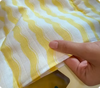 A close-up of a yellow and white muslin fabric with wavy stripes. A person’s hand is gently holding the edge of the fabric, showcasing its texture. The yellow stripes add a playful and soft design to the fabric, suitable for baby products like swaddles or blankets. The light fabric appears soft and breathable, emphasizing comfort and care for infants.